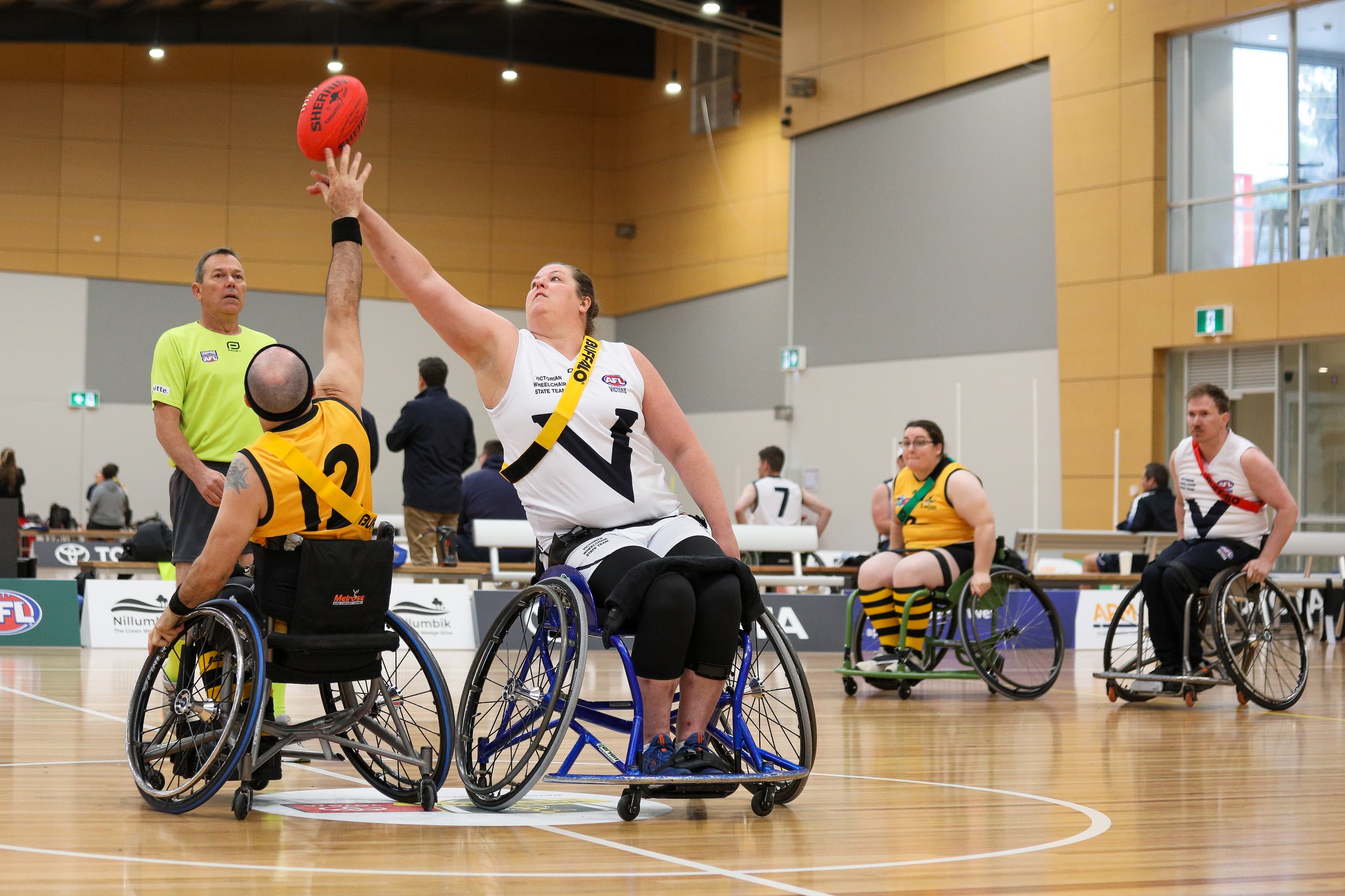 A group of wheelchair AFL players on an indoor court. Two players are reaching up for the football as the referee looks on.