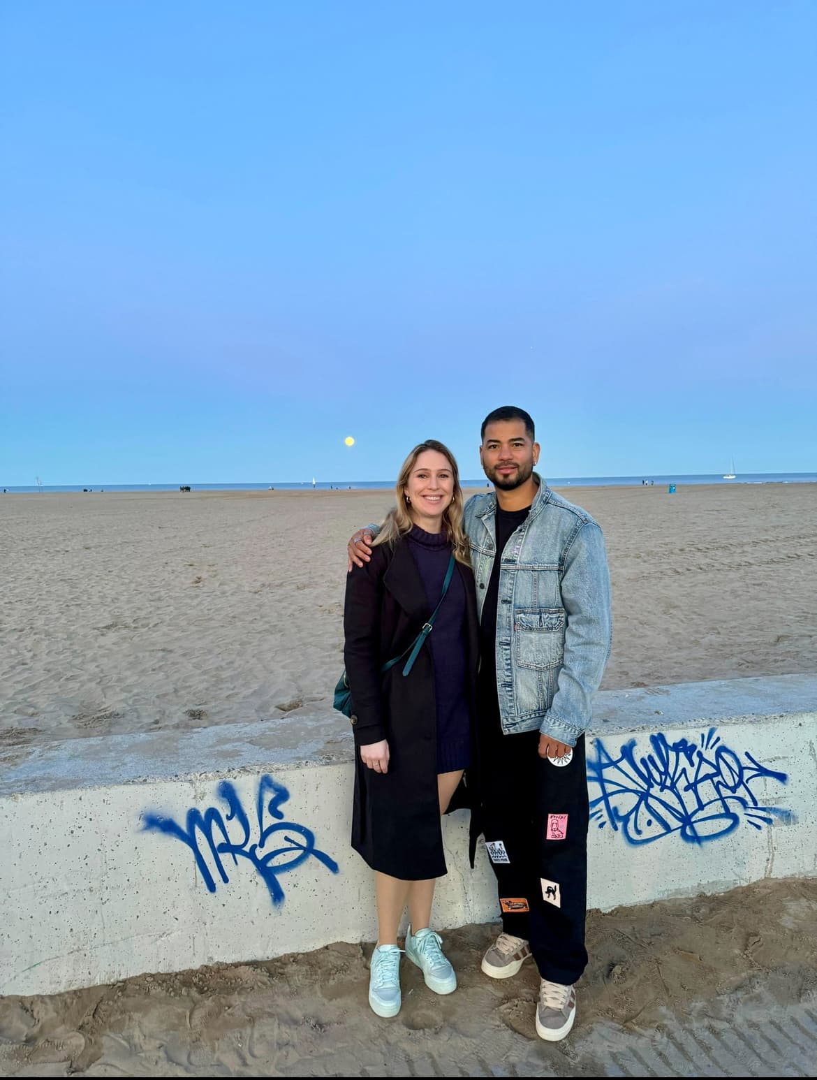 Emily and her partner standing in front of a beach.