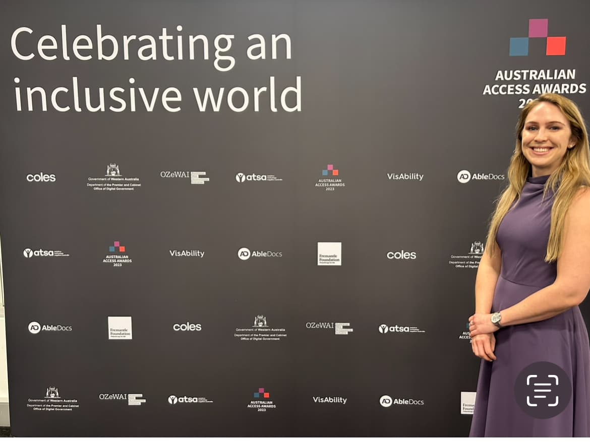 A photo of Emily standing in front of a banner. She has light coloured hair and is wearing a purple dress. On the banner are the words ‘celebrating an inclusive world’ in white text on a dark background.