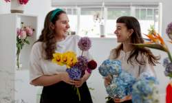 Two young women are standing in a florist. They are looking at each and smiling. They are both wrapping bunches of flowers.