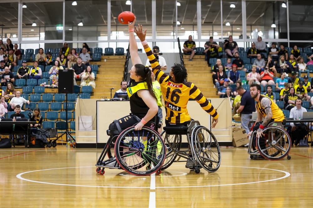 Two people in sportschairs are playing AFL on an indoor court. They are back to back and both reaching for the football above them.