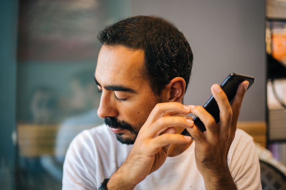 A man is holding his phone up to his ear and listening to it. He appears to be using a screen reader. 