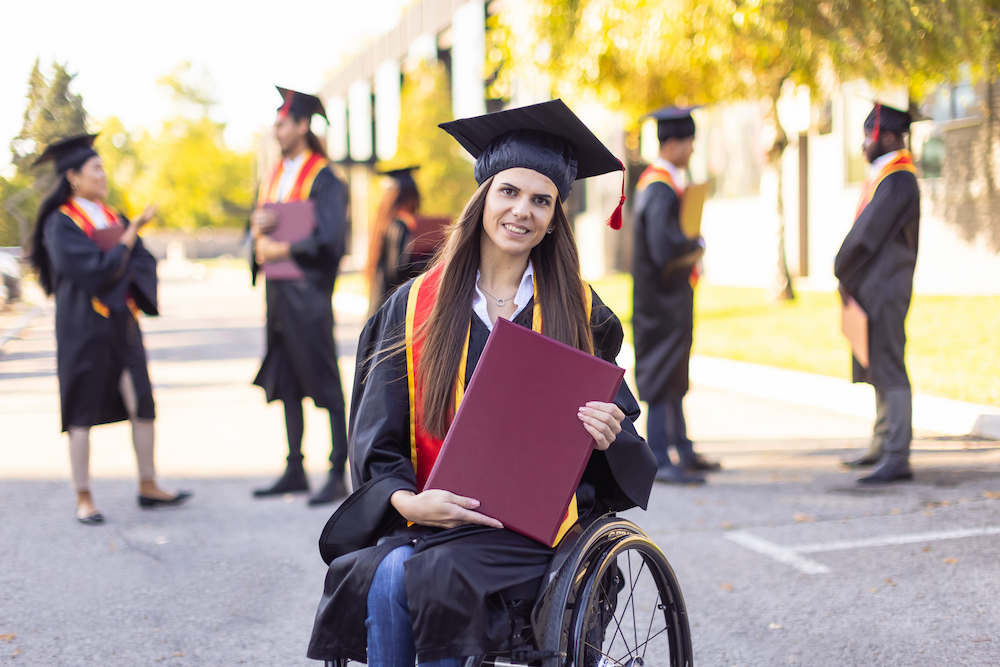 A smiling girl in a wheelchair is wearing a graduation cap and gown and holding a diploma. She is outside.