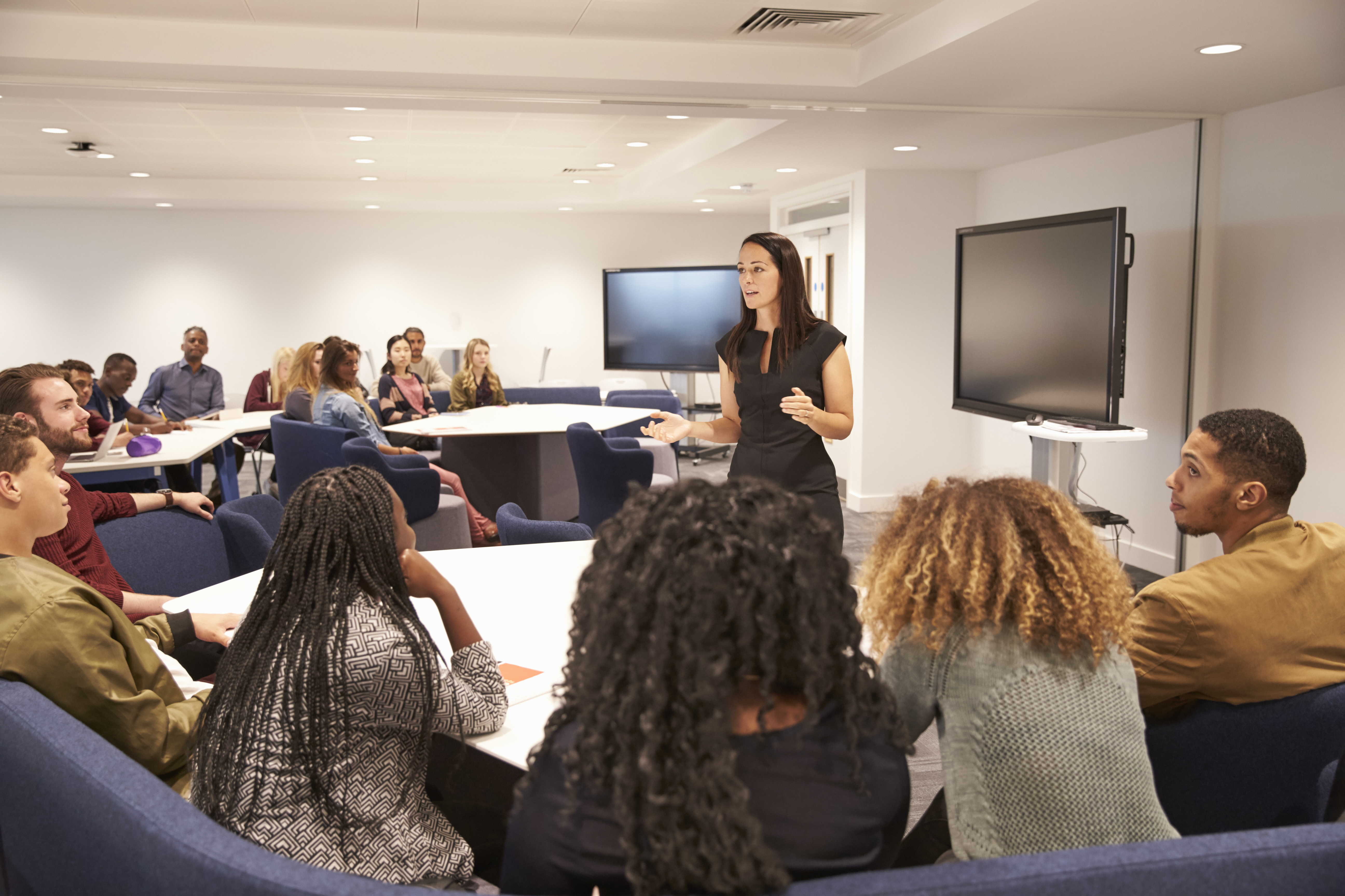 A woman is standing in the centre of a room with screens behind her. She is gesturing and talking. There are people sitting at tables around her listening. 