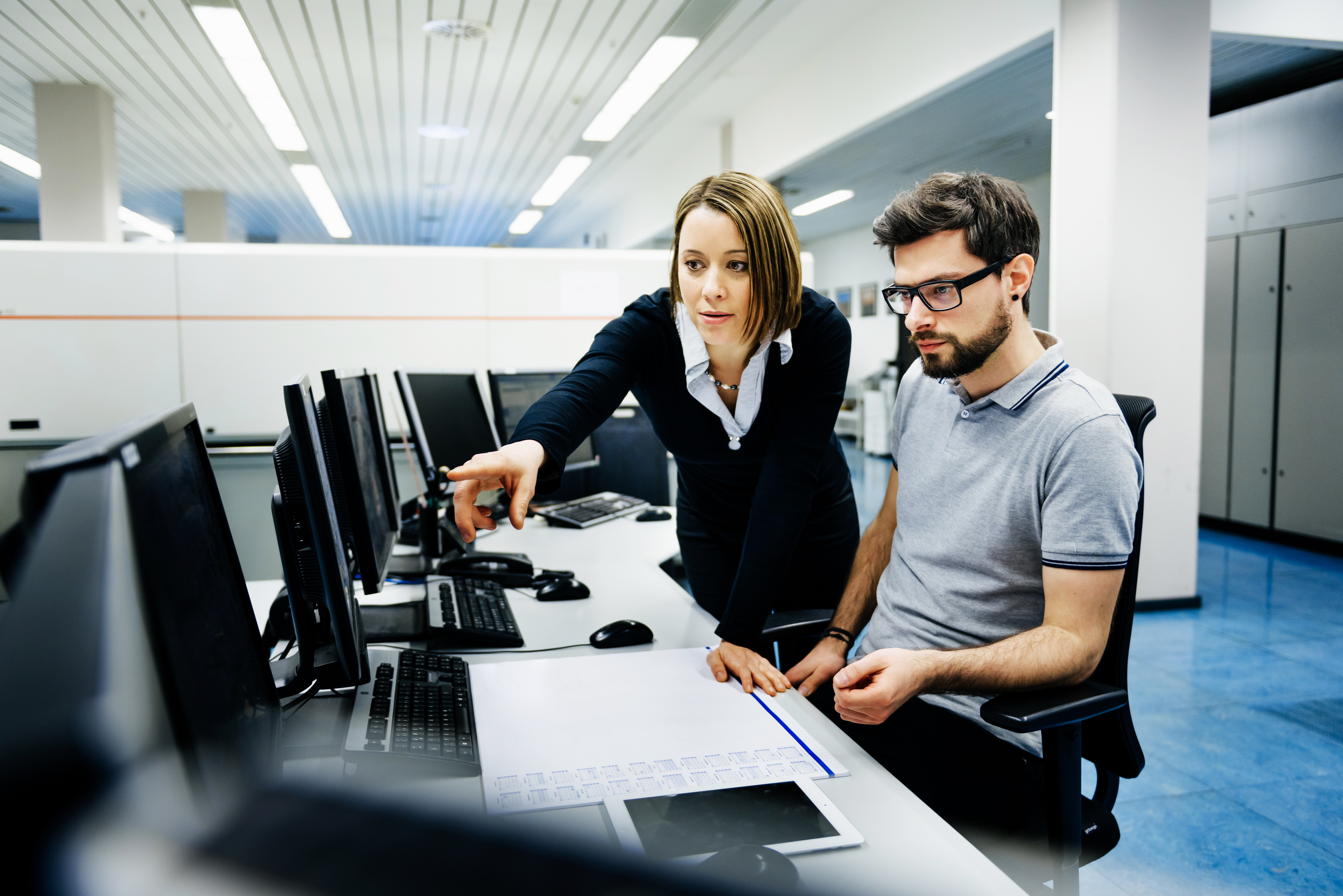 A man and a woman are looking at a computer together. The woman is pointing at the screen and explaining something. They are in a professional workplace.