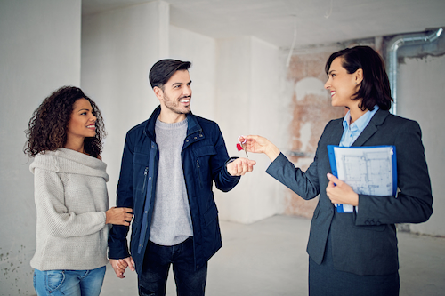 A real estate agent is handing a couple a house key and holding some paperwork. They are all standing inside the house and smiling. 