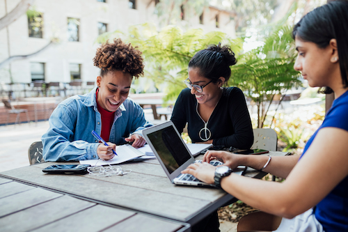 3 young women are sitting at an outdoor table studying. One of them is typing on a laptop. The other two are smiling and writing notes.